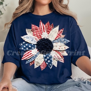Può includere: T-shirt blu navy con un grande motivo a girasole. I petali sono in tessuto con motivi rossi, bianchi e blu, tra cui stelle e strisce. Il centro del fiore è un pompon blu scuro.