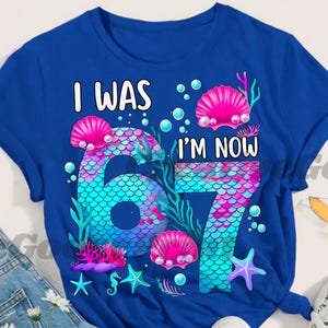 Puede incluir: Camiseta azul real con el texto "I WAS I'M NOW 67". Los n&uacute;meros est&aacute;n dise&ntilde;ados con un patr&oacute;n de escamas de sirena en tonos de azul, rosa y morado. El dise&ntilde;o incluye conchas marinas, burbujas y estrellas de mar.