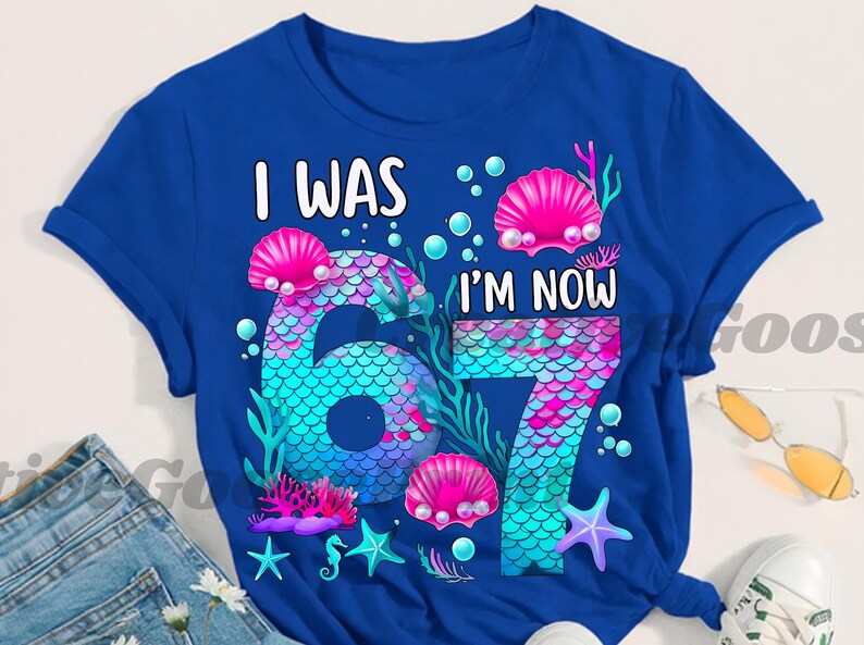 Puede incluir: Camiseta azul real con el texto "I WAS I'M NOW 67". Los n&uacute;meros est&aacute;n dise&ntilde;ados con un patr&oacute;n de escamas de sirena en tonos de azul, rosa y morado. El dise&ntilde;o incluye conchas marinas, burbujas y estrellas de mar.