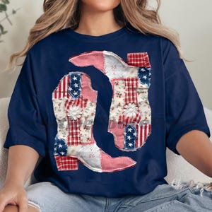 May include: Navy blue t-shirt featuring a patchwork design of two cowboy boots. The boots are decorated with red, white, and blue patterns, including stars and stripes, and floral prints. White flower accents are placed on the boots.