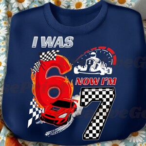 May include: Navy blue sweatshirt with a racing theme. Features the text "I WAS 6 NOW I'M 7" with a large red number 6 engulfed in flames, a red race car, and a checkered flag design. A speedometer and karting graphic are also included.
