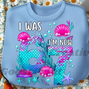 Puede incluir: Sudadera azul claro con el texto "I WAS 6, I'M NOW 7". El dise&ntilde;o presenta un tema de sirena con los n&uacute;meros 6 y 7 en un patr&oacute;n de escamas, conchas rosas y elementos acu&aacute;ticos.