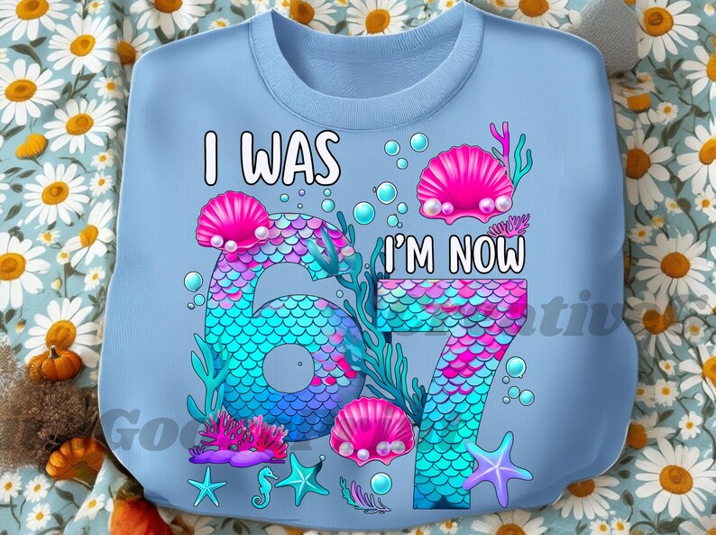 Puede incluir: Sudadera azul claro con el texto "I WAS 6, I'M NOW 7". El dise&ntilde;o presenta un tema de sirena con los n&uacute;meros 6 y 7 en un patr&oacute;n de escamas, conchas rosas y elementos acu&aacute;ticos.