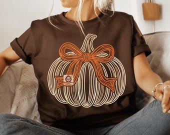 May include: A dark brown short-sleeved t-shirt featuring a large graphic of a white-lined pumpkin adorned with a prominent orange bow. The design includes a small square logo with a stylized owl character positioned on the pumpkin graphic.