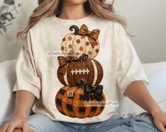 May include: A cream-colored t-shirt featuring a graphic of three stacked pumpkins. The top pumpkin is cream with brown polka dots and a brown bow. The middle pumpkin is brown with white football laces. The bottom pumpkin has a black and orange buffalo plaid pattern with a black bow.