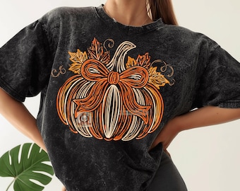 May include: A black mineral-washed short-sleeve t-shirt featuring a large graphic print of an orange and white pumpkin adorned with a prominent orange bow and stylized autumn leaves, with thin swirling vine details surrounding the pumpkin.