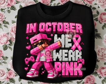 May include: A black sweatshirt featuring a graphic of a pixelated character in a pink outfit performing a dab pose. The text reads IN OCTOBER WE WEAR PINK in block letters. The design includes a pink awareness ribbon and small pink game controllers.