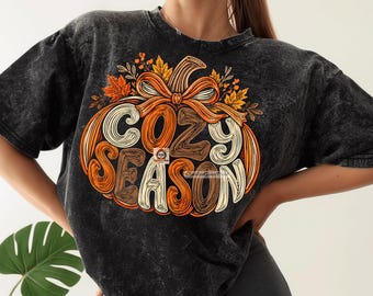 May include: A black acid-wash t-shirt featuring a large graphic of an orange pumpkin with a bow on top. The pumpkin is decorated with autumn leaves and the text COZY SEASON in a stylized font. The design incorporates shades of orange, brown, and cream.