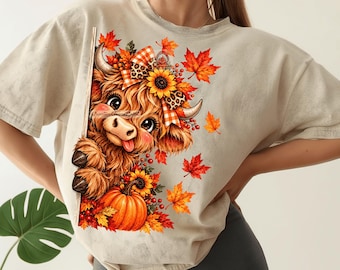 May include: A beige t-shirt featuring a graphic print of a fluffy brown Highland cow with a plaid and leopard print bow, sunflowers, red berries, and orange maple leaves. A bright orange pumpkin sits at the base of the cow illustration.