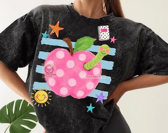 May include: A black acid-wash t-shirt featuring a graphic of a pink polka-dot apple with a green worm wearing pink glasses. The design includes light blue horizontal stripes, a yellow smiling sun, colorful stars, and a small white notebook.