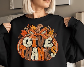 May include: A black crewneck sweatshirt featuring a large graphic of a stylized pumpkin with an orange bow on top. The pumpkin is filled with the text GIVE THANKS in a decorative, multi-colored font, surrounded by autumn leaves and branches.