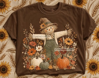May include: A brown t-shirt featuring a graphic of a scarecrow wearing a hat and overalls, surrounded by autumn flowers and three pumpkins in white, green, and orange. The design includes the text CreativeGoosePrint in two locations.