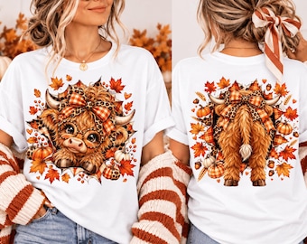 May include: A white t-shirt featuring a graphic of a fluffy Highland cow wearing glasses and a plaid bow, surrounded by autumn leaves and pumpkins. The front shows the cow's face, while the back displays the cow's rear with a matching plaid bow.
