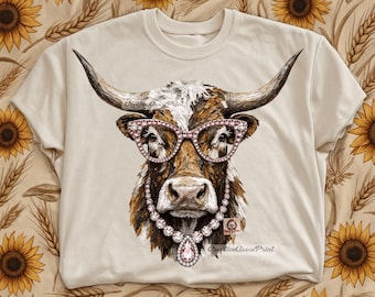 May include: A cream-colored t-shirt featuring a graphic illustration of a longhorn cow wearing pink rhinestone cat-eye glasses and a matching rhinestone necklace with a large teardrop pendant. The cow has brown and white fur.