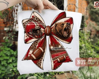 May include: A white t-shirt featuring a large graphic of a bow. The bow design incorporates a red football helmet, a roaring brown bear, two brown footballs, a leopard print pattern, and a stadium scene with red and white colors.