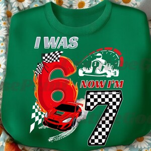 May include: Green sweatshirt with a racing theme. Features the text "I WAS 6 NOW I'M 7" with a large red number 6 on fire, a red race car, and a checkered flag. Includes a graphic of a go-kart.