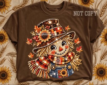 May include: A brown t-shirt featuring a graphic of a scarecrow wearing a plaid hat adorned with a sunflower and autumn leaves. The scarecrow has a stitched smile, a triangular nose, and wears a plaid scarf and blue overalls with a sunflower.