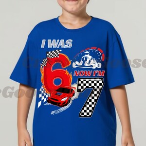 May include: Royal blue t-shirt with a racing design. The shirt displays the text "I WAS 6, NOW I'M 7" with a large red number 6 on fire, a red race car, and a checkered flag. A speedometer and a kart are also visible.