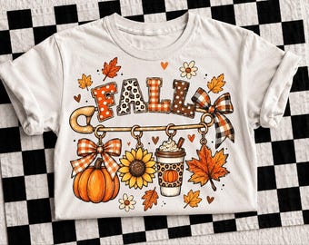 May include: A white t-shirt featuring a large graphic of a gold safety pin with the word FALL spelled out in patterned letters. Hanging from the pin are a pumpkin with a plaid bow, a sunflower, a coffee cup with a pumpkin print, and a maple leaf.