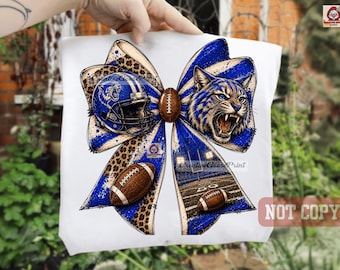 May include: A white t-shirt featuring a large graphic of a bow with blue and leopard print patterns. The design includes a football helmet, a snarling wildcat, two brown footballs, and a football field scene with the text CreativeGoosePrint.