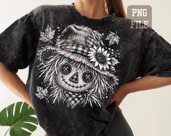 May include: A black acid-wash t-shirt featuring a white graphic print of a scarecrow with button eyes, a stitched mouth, and a plaid hat adorned with a sunflower. The design includes scattered maple leaves and a small plaid neck scarf detail.