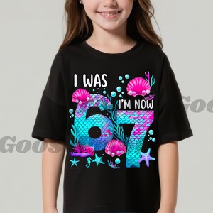 Puede incluir: Camiseta negra con un dise&ntilde;o de sirena. El gr&aacute;fico presenta los n&uacute;meros "6" y "7" en un patr&oacute;n de escamas de sirena, rodeados de conchas, estrellas de mar y el texto "I WAS 6, I'M NOW 7".
