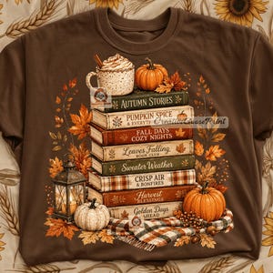 Op de afbeelding: Een bruin t-shirt met een afbeelding van een stapel boeken met de titels Autumn Stories, Pumpkin Spice, Fall Days, Leaves Falling, Sweater Weather, Crisp Air en Harvest Blessings, bekroond met een mok en pompoenen, omringd door herfstbladeren.
