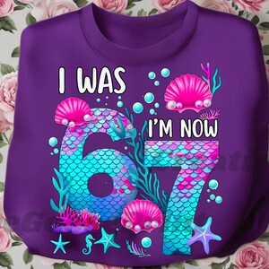 Puede incluir: Una sudadera morada con el texto "I WAS 67 I'M NOW 67" en blanco. Los n&uacute;meros est&aacute;n dise&ntilde;ados con un patr&oacute;n de escamas de sirena en tonos azules y turquesas. Conchas marinas rosas y estrellas de mar decoran el dise&ntilde;o.