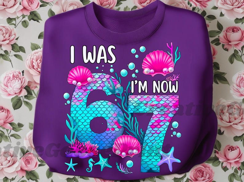 Puede incluir: Una sudadera morada con el texto "I WAS 67 I'M NOW 67" en blanco. Los n&uacute;meros est&aacute;n dise&ntilde;ados con un patr&oacute;n de escamas de sirena en tonos azules y turquesas. Conchas marinas rosas y estrellas de mar decoran el dise&ntilde;o.