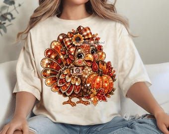 May include: A cream-colored t-shirt featuring a graphic of a turkey with a mosaic-style pattern in shades of orange, brown, and gold. The turkey wears a plaid bow and is surrounded by autumn leaves and a pumpkin, all rendered in a sparkling effect.