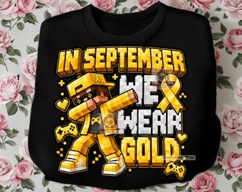 May include: A black sweatshirt featuring a pixelated yellow character performing a dabbing pose. The graphic includes a yellow awareness ribbon and the text IN SEPTEMBER WE WEAR GOLD in a blocky, pixel-style font, accented by small yellow hearts and game controllers.