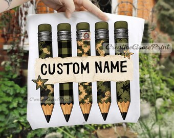 May include: A white t-shirt featuring a graphic of five pencils with various green camouflage and plaid patterns. A horizontal, torn-paper style banner across the center displays the text CUSTOM NAME in black, flanked by two small orange stars.