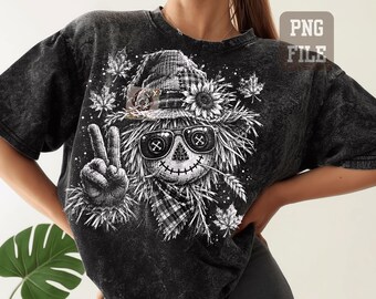 May include: A black acid-wash t-shirt featuring a white graphic of a scarecrow wearing a plaid hat and sunglasses, making a peace sign. The design includes sunflowers, maple leaves, and a plaid neckerchief, with a stitched mouth and button eyes.