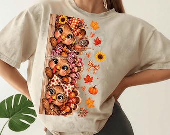 May include: A beige short-sleeve t-shirt featuring a graphic print of three turkeys stacked vertically behind a wooden fence post. The turkeys wear patterned bows and glasses, surrounded by autumn leaves, sunflowers, and orange pumpkins.