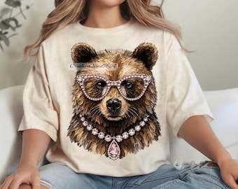May include: A cream-colored t-shirt featuring a graphic print of a brown bear wearing pink rhinestone-encrusted cat-eye glasses and a matching rhinestone necklace with a large pink teardrop pendant. The text CreativeGoosePrint is visible.