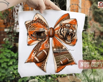 May include: A white t-shirt featuring a large graphic of an orange and black bow. The design includes a football helmet, a roaring tiger, two footballs, and a stadium field scene, all accented with leopard print patterns and orange splatters.