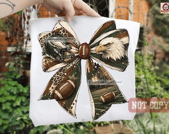 May include: A white t-shirt featuring a large graphic of a bow. The bow design includes a football helmet, a bald eagle, two footballs, and a football field at night. The pattern incorporates leopard print, dark green, and brown color accents.