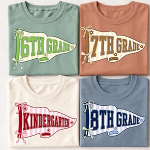 May include: Four crew neck t-shirts in sage green, terracotta, cream, and slate blue. Each shirt features a pennant graphic with a striped background, ribbon bows, and small flowers. The text reads 6TH GRADE, 7TH GRADE, KINDERGARTEN, and 8TH GRADE.