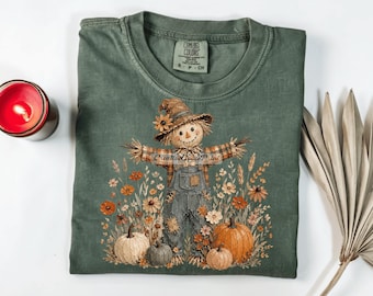 May include: A sage green t-shirt featuring a graphic of a scarecrow wearing a hat and overalls, surrounded by pumpkins and wildflowers. The shirt has a Comfort Colors label at the neck and a watermark reading CreativeGoodPrint across the center.