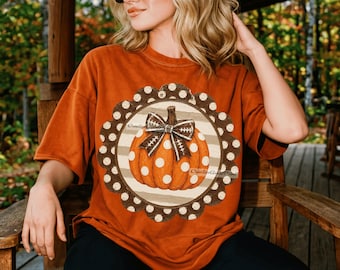 May include: An orange short-sleeved t-shirt featuring a central graphic of an orange pumpkin with white polka dots. The pumpkin is centered within a brown scalloped circle frame with white dots and a brown and white football-patterned bow.