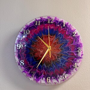 May include: Round wall clock with a colorful abstract design in shades of purple, blue, and red. The clock face features silver numerals and gold hands. The clock is mounted on a light gray wall.
