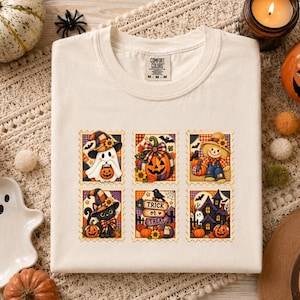 Puede incluir: Una camiseta de color crema con un estampado gráfico de seis diseños rectangulares de sellos postales. Las ilustraciones incluyen un fantasma, una calabaza, un espantapájaros, un gato negro, un letrero que dice Trick or Treat y una casa encantada.