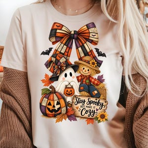 以下が含まれることがあります： ベージュのTシャツで、パッチワーク柄の幽霊、かかし、カボチャのグラフィックプリントが施されています。上部には大きなチェック柄のリボンがあり、下部には「Stay Spooky but make it cozy」という文字が書かれた木製の看板があります。