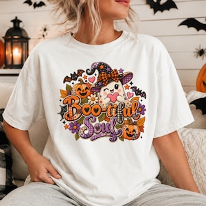 Puede incluir: Una camiseta blanca de manga corta con un estampado gráfico colorido de un fantasma de dibujos animados con sombrero de bruja, rodeado de calabazas, murciélagos, flores y corazones. El texto "Boo-tiful Soul" está impreso en una fuente estilizada.