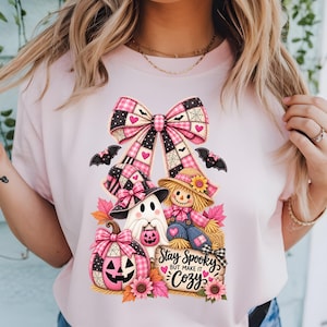 Puede incluir: Una camiseta rosa claro con un estampado gráfico de un gran lazo a cuadros rosa y negro sobre un fantasma con sombrero de bruja, un espantapájaros y una calabaza. El diseño incluye murciélagos, hojas de otoño y el texto Stay Spooky BUT MAKE IT Cozy.