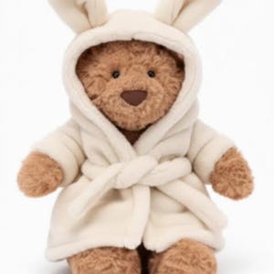 May include: A plush teddy bear wearing a cream-colored bunny-eared robe. The bear has brown fur, a brown nose, and black eyes. The robe is tied at the waist with a matching belt. The text "PLUSHIECHARMZ" is at the bottom.