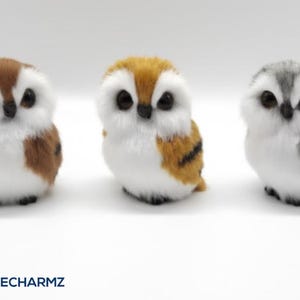 May include: Three plush owl figurines in brown, orange, and gray with white bellies and large black eyes. The owls are small and fluffy, with a realistic appearance. The brand name "PLUSHIECHARMZ" is at the bottom.