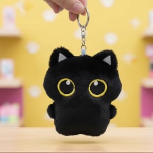 Puede incluir: Un llavero de gato de peluche negro con grandes ojos con borde amarillo y pequeños acentos triangulares blancos en las orejas. El llavero está sostenido por una mano sobre un fondo amarillo. La palabra "PLUSHIECHARMZ" está en la parte inferior.