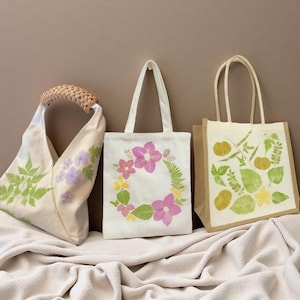 May include: Three tote bags with floral and leaf designs. One bag has a woven handle and floral prints in green and purple. Another features a pink floral wreath on a white background. The third bag has a natural jute base with leaf prints.