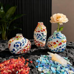 May include: Three mosaic vases in various shapes and sizes, adorned with colorful glass pieces. One vase holds a single flower. Scattered around are colorful glass pieces and a tube of adhesive.
